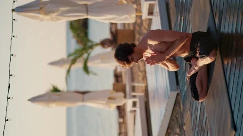 A guy in black shorts sits on a mat and does yoga on the beach in the morning 스톡 동영상 249605985