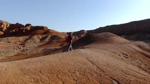 A guy with a camera walks along a hill against the backdrop of red mountains Stock Footage 261850223