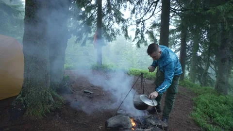 A guy cooks food in a cauldron on a fire on a camping trip with a tent. Stock Footage 214323256