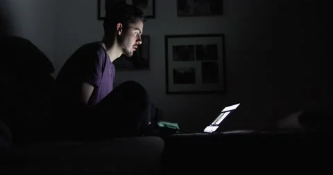 Guy in a dark room drinking tea on the sofa looks at the screen of the laptop Stock Footage 130489565