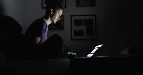 Guy in a dark room sits on the sofa and looks at the screen of the laptop Stock Footage 130489604