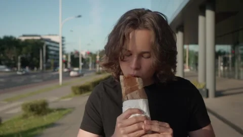 A guy is eating a shawarma, taking slow bites as he strolls down a city street. Stock Footage 296768136