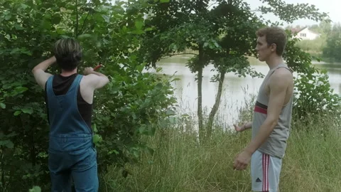 A guy in a farm jumpsuit walks up to a tree with a guy in a tracksuit Stock Footage 197481542