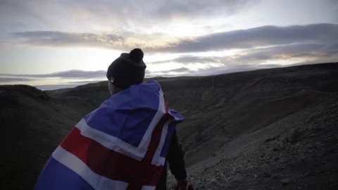 A guy with the flag of Iceland stands on the edge of a cliff and looks at the Stock Footage 130450690