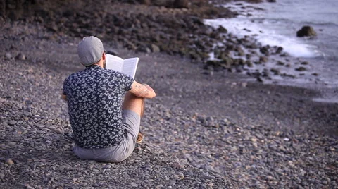 A guy with flat cap reading a book sitting on the beach Stock Footage 58212213