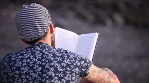 A guy with flat cap reading a book sitting on the beach Stock-Footage 58214196