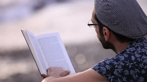 A guy with flat cap reading a book sitting on the gravel beach Stock Footage 58256764