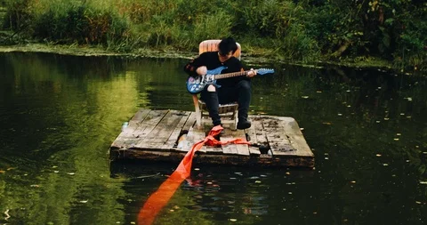 Guy fun playing guitar sitting on a raft in the middle of the pond Stock Footage 114520410