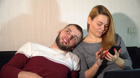 The guy with the girl is sitting on the couch, the guy is bored, and the girl is 스톡 동영상 86148585