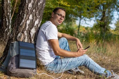 A guy in glasses sits by a tree and charges his phone with a solar panel Stock Photos