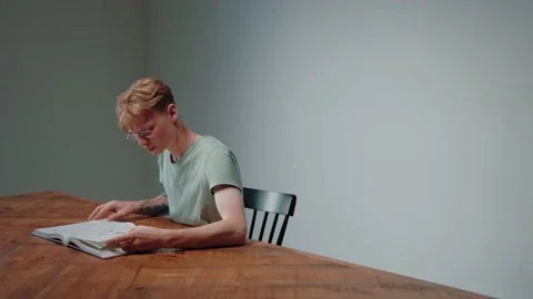 Guy in glasses studying by reading a textbook, sitting in empty grey room Vídeo Stock 164372402
