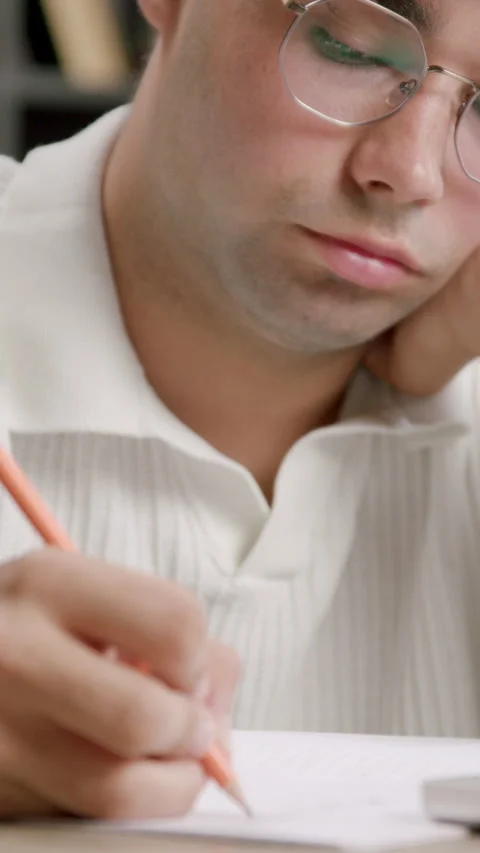 Guy in glasses writes propping up head with hand at desk Stock Footage 313926137