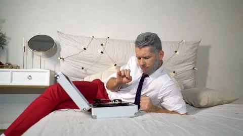 A guy with a gray beard in red trousers is lying in bed and listening to music Video stock 167651210