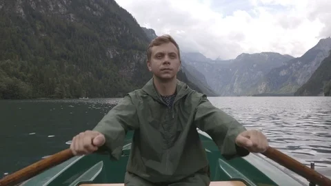 Guy in the green raincoat is rowing on the boat  on the mountain lake Stock Footage 83626975