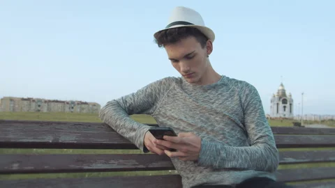 Guy in a hat and with an earring in his ear, who sits on an old city bench with 스톡 동영상 166979580