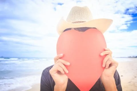 Guy in Hat Hides Face behind Hand-made Red Heart on Beach Fotos Stock