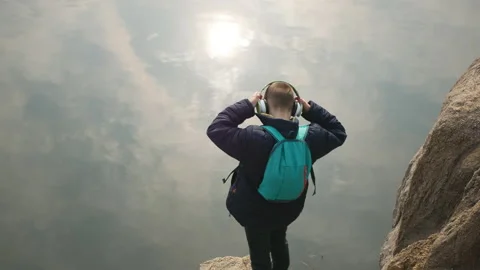 A guy with headphones and a backpack is standing on the shore of the lake. Stock Footage 105286107
