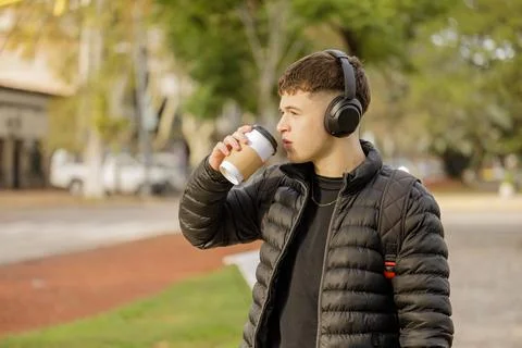 Guy with headphones drinks coffee while walking through a public park with .. Stock Photos