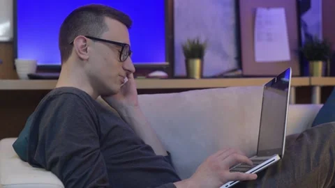 The guy at home is sitting on the couch with a laptop, and talking on the pho Stock Footage 130217027