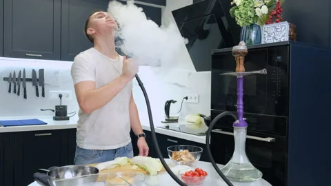 Guy in Kitchen at Table with Products. Smokes Hookah. Deeply Draws In Smoke Stock Footage 147003636