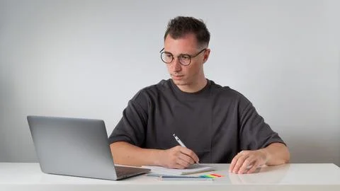 A guy at a laptop makes notes in a notebook at the workplace - work and study Stock Photos