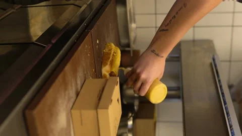 Guy In A Leather Apron Is Preparing A Hot Dog. Chef In Gloves Pours Mustard On Stock Footage 196200118