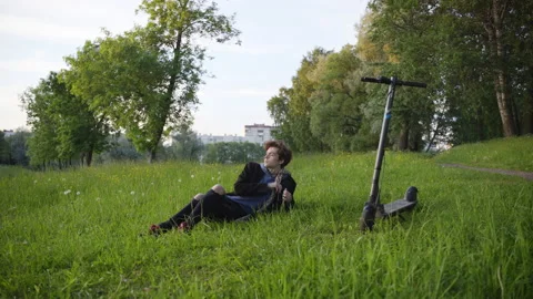 A guy lies on the grass with a phone, gets up, leaves on an electric scooter. Stock-Footage 201075603