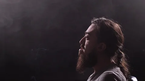 A guy with a long beard and oblique smokes a cigarette on a black background Stock Footage 105280305