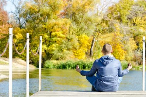 Guy in the lotus pose sits on the dock. Autumn, sunny. Back view Stock Photos