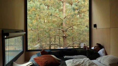 Guy is lying in bed, dressed, and looking at his smartphone view of the window Stock Footage 289437757