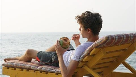 Guy lying on a chaise longue sips coconut water through a straw Stock Photos