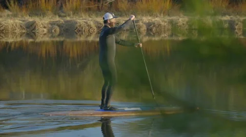 Guy paddle boards away down river in side view Stock Footage 63386126