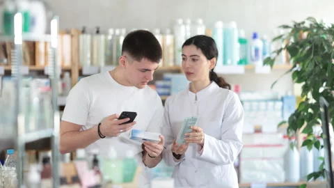 Guy in pharmacy scans QR code on antifungal ointment, pharmacist in background Stock Footage 316580397