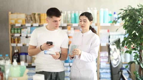Guy in pharmacy scans QR code on antifungal ointment, pharmacist in background Stock Footage 330039922