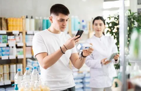 Guy in pharmacy scans QR code on antifungal ointment, pharmacist in background 스톡 사진