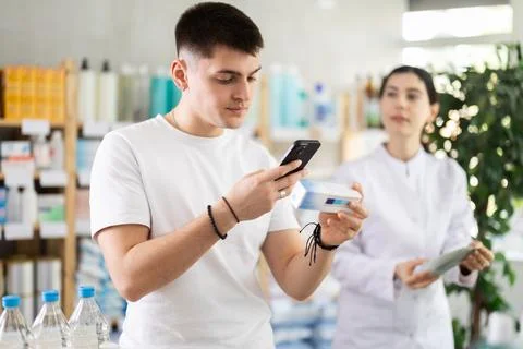 Guy in pharmacy scans QR code on antifungal ointment, pharmacist in background Stock Photos
