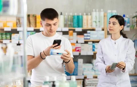 Guy in pharmacy scans QR code on antifungal ointment, pharmacist in background Fotos de archivo