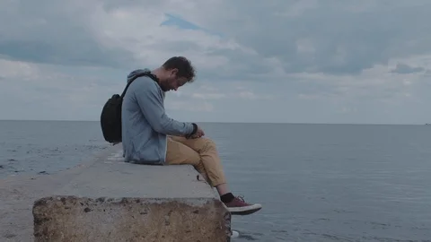 Guy a photographer with a beard sits on a pier in the sea and sets up his camera Stock Footage 115480416