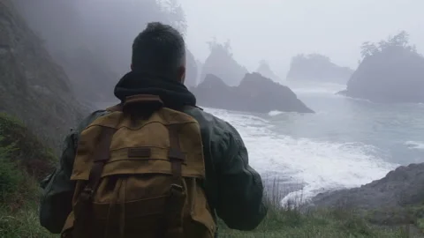 Guy in pilot jacket traveling by green mountain to rocky coastline, Back view Video stock 166932815