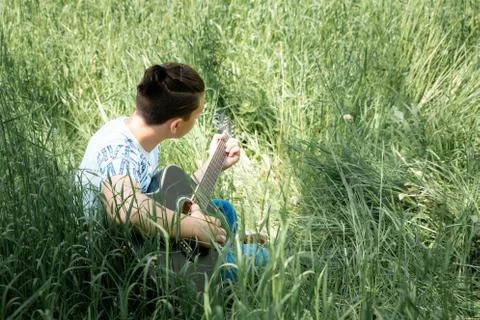 A guy playing a guitar sitting in the back of a green grass Stock Photos