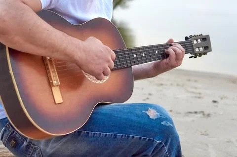 A guy plays an acoustic six-string guitar on a sandy beach by a body of water Stock Photos