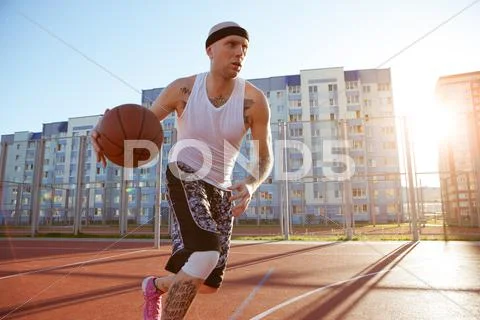 Guy Plays Basketball On A Red Court In Sunny Weather The Background Of Houses