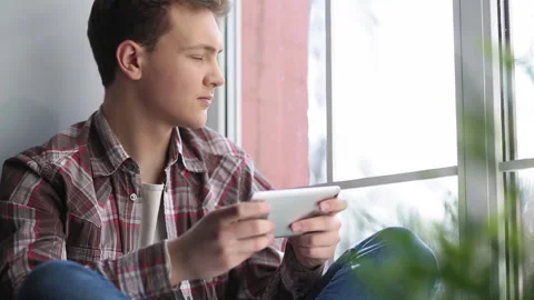 The guy plays on the smartphone while sitting on the windowsill by the window Stock Footage 130538413
