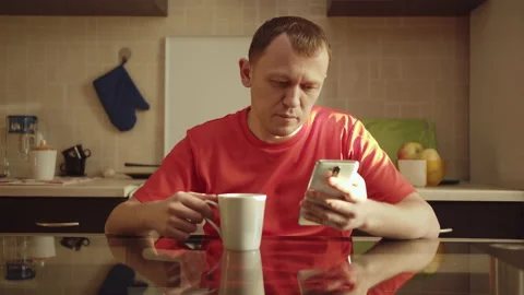 The guy reads the message in his phone while sitting in the kitchen drinking tea Stock Footage 130873997