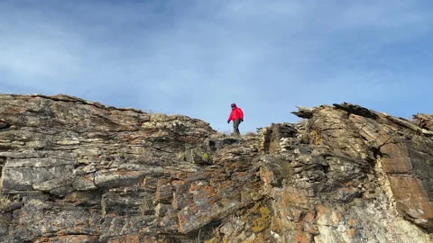 A guy in a red jacket walks along the edge of a rocky mountain Stock Footage 151064020