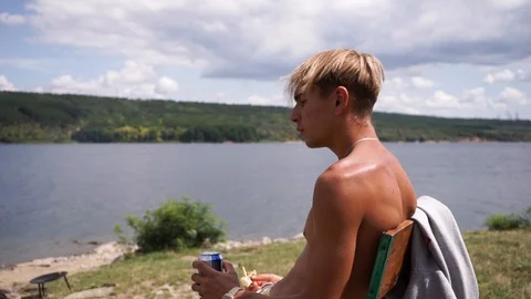 The guy is resting on the river bank, drinking cool fresh beer, hot summer day Stock Footage 114630991