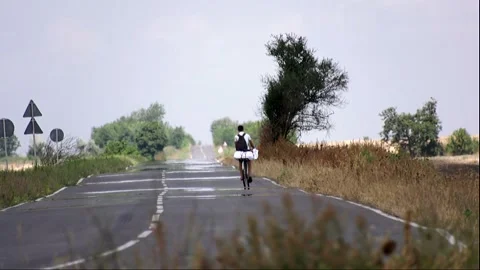 A guy is riding a bicycle along a long road between fields in summer Stock Footage 324766771