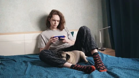 A guy with a serious look sits in bed with a phone, a dog sleeps next to him 動画素材 196813423