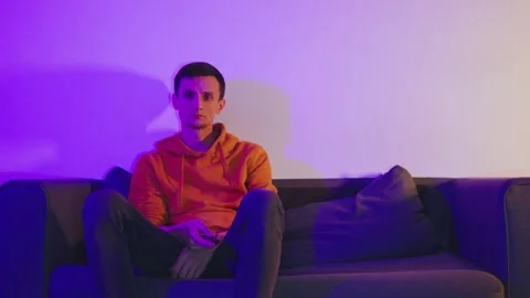 A guy sits on the couch with a remote in hand, flipping through TV channels Stock Footage 297562567