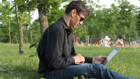 Guy sits on the grass stretching his leg with a laptop and typing Stock Footage 135238224